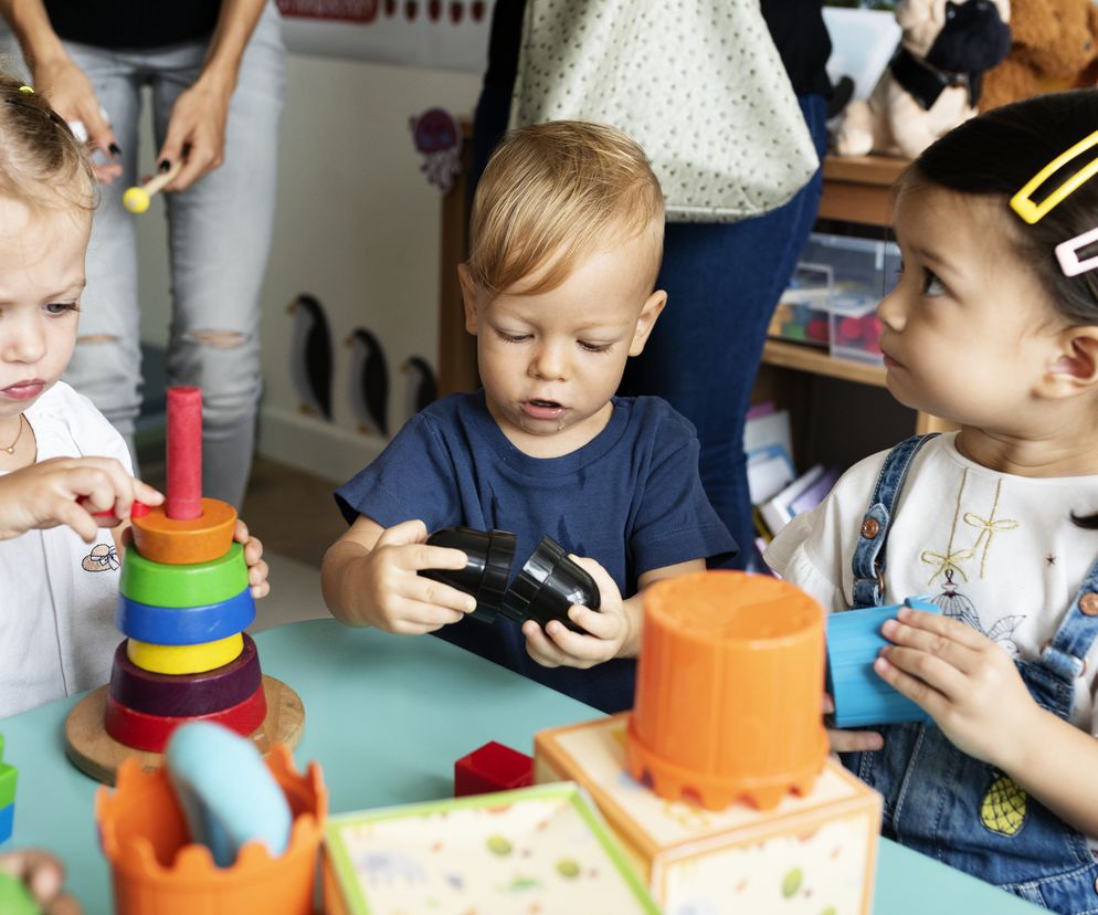 Troje małych dzieci w wieku przedszkolnym, prawdopodobnie stock, w trakcie zabawy edukacyjnymi zabawkami konstrukcyjnymi przy zielonym stoliku. W tle widać postacie dorosłych. Na Poradniku Zdrowie znajdziesz informacje o nowych zasadach w przedszkolach i wytycznych GIS.