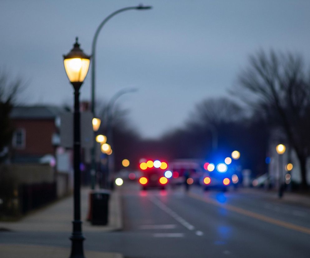 W centrum kadru dominują dwa rzędy ulicznych lamp, z których jedna, na pierwszym planie po lewej stronie, świeci intensywnym, ciepłym, żółtym światłem, ukazując delikatne detale czarnej, metalowej obudowy. Droga asfaltowa rozciąga się w głąb obrazu, rozświetlona zamazanymi światłami awaryjnymi pojazdów – czerwonymi i niebieskimi po prawej stronie oraz żółtymi i czerwonymi po lewej. W tle, niewyraźne sylwetki budynków i drzew rysują się pod pochmurnym, ciemnoszarym niebem.