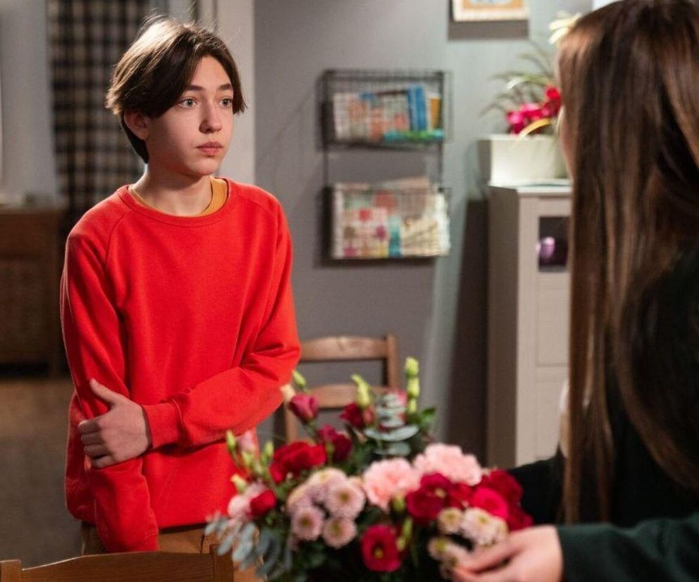 Młody Emil (Artem Malaszczuk) w czerwonej bluzie z długimi rękawami, stojący w pokoju, z zmartwioną miną, patrzy na Agatę (Natalia Zambrzycka), która układa kwiaty, co zapowiada tragiczną wiadomość o zniknięciu Ignacego z serialu Barwy szczęścia na Super Seriale.