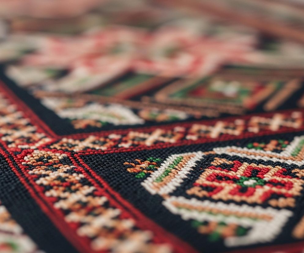 Zbliżenie na fragment tkaniny o wyraźnym wzorze ludowym. Dominują geometryczne motywy w odcieniach czerwieni, zieleni, beżu, czerni i bieli, ułożone w symetryczne pasy i kwadraty. Kolory są nasycone, a faktura tkaniny, widoczna dzięki ostrości w pierwszym planie, wskazuje na haft lub tkactwo. W głębi obrazu widoczne są rozmyte, większe kształty w pastelowych odcieniach różu, które mogą być częścią dalszego wzoru lub innego elementu tła.