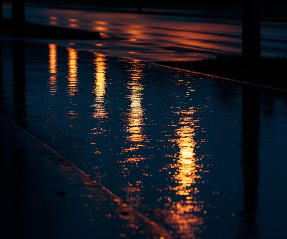 Mokry asfalt lub inna ciemna, płaska powierzchnia odbija intensywne, pomarańczowo-złote światła, tworzące podłużne smugi i rozproszone plamy na granatowo-czarnej tafli. Po prawej stronie widać ciemne sylwetki słupów lub drzew, których odbicia również są widoczne w mokrej nawierzchni. Horyzont w górnej części obrazu jest zamazany, a odbite światło rozciąga się w głąb perspektywy.