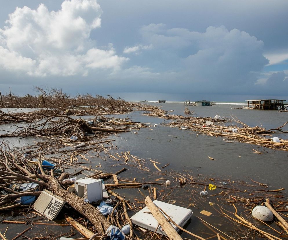 Akwen wodny o ciemnej, wzburzonej powierzchni wypełniony jest dryfującymi gałęziami, kawałkami drewna, krzakami oraz gruzem i śmieciami, w tym plastikowymi butelkami, styropianowymi elementami, kawałkami metalu i fragmentami urządzeń gospodarstwa domowego. Po lewej stronie, bliżej obserwatora, w wodzie leżą zniszczone sprzęty, takie jak biała szafka i szary klimatyzator, otoczone plątaniną gałęzi. Na dalszym planie, na wysokości horyzontu, widać kilka zniszczonych budynków, w tym jeden większy, drewniany, osadzony na palach, częściowo zanurzony w wodzie. Niebo jest częściowo pokryte dużymi, białymi chmurami, a w prawym górnym rogu widać fragmenty błękitnego nieba.