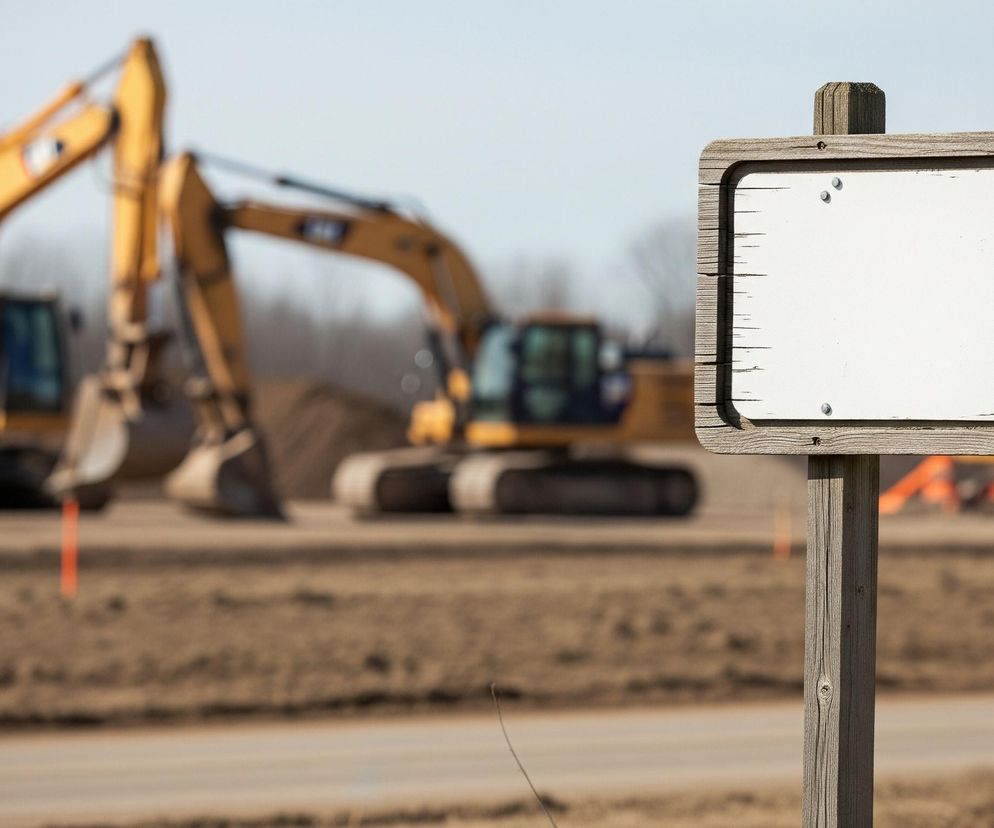 Pusta drewniana tablica informacyjna na tle rozmytego placu budowy z żółtymi koparkami i piaszczystym terenem, symbolizująca kontrowersje wokół działek pod CPK. Więcej o aferze przeczytasz na portalu Super Biznes.