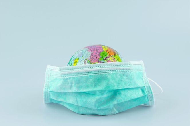 Globus w zielonej maseczce ochronnej na jasnym tle, symbolizujący pandemię koronawirusa i globalne restrykcje, w tym zakaz ruchu lotniczego i plany wakacyjne. Więcej o wpływie COVID-19 na podróże i zdrowie przeczytasz na Poradnik Zdrowie.
