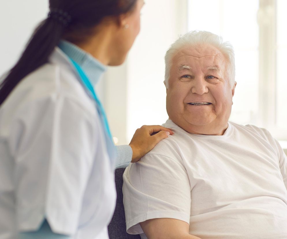 Uśmiechnięty starszy mężczyzna z siwymi włosami, w białej koszulce, siedzi na kanapie, a obok niego stoi opiekunka w białym kitlu i turkusowym podkoszulku, trzymając dłoń na jego ramieniu. Artykuł na Super Biznes informuje o podwyżce świadczeń pielęgnacyjnych w 2026 roku.