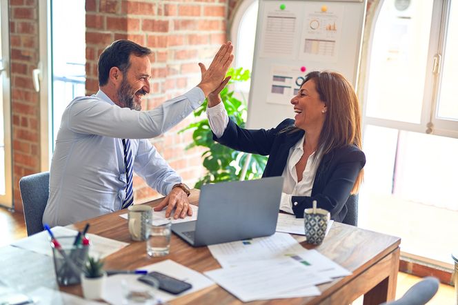 Uśmiechnięci przedsiębiorcy, mężczyzna i kobieta, wymieniają high-five przy biurku z laptopem i dokumentami. Symbolizują współpracę i sukces w biznesie, omawiając ważne kwestie dla firm, o czym można przeczytać na Super Biznes.