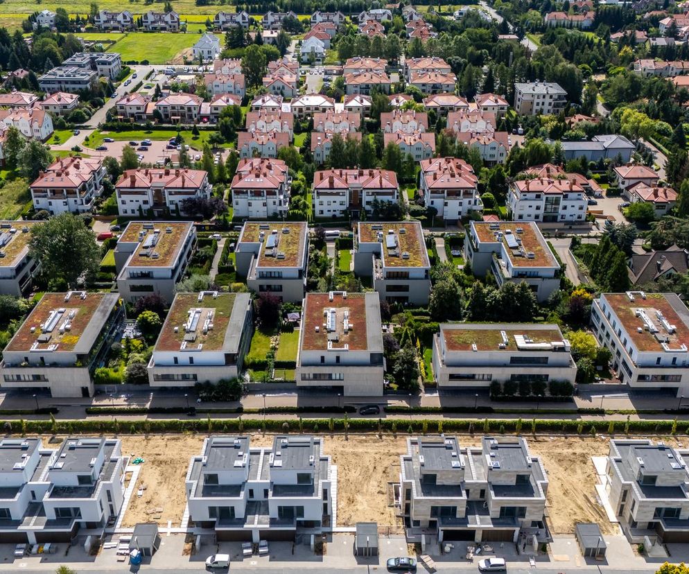 Ujęcie z lotu ptaka przedstawia zurbanizowane osiedle łanowe na warszawskich Zawadach, ukazujące różnorodność zabudowy – od nowo budowanych domów jednorodzinnych, przez modernistyczne bloki z zielonymi dachami, po tradycyjne domy z czerwonymi dachami, co jest typowe dla dynamicznych zmian architektonicznych, o których możesz przeczytać na Architektura Murator Plus.