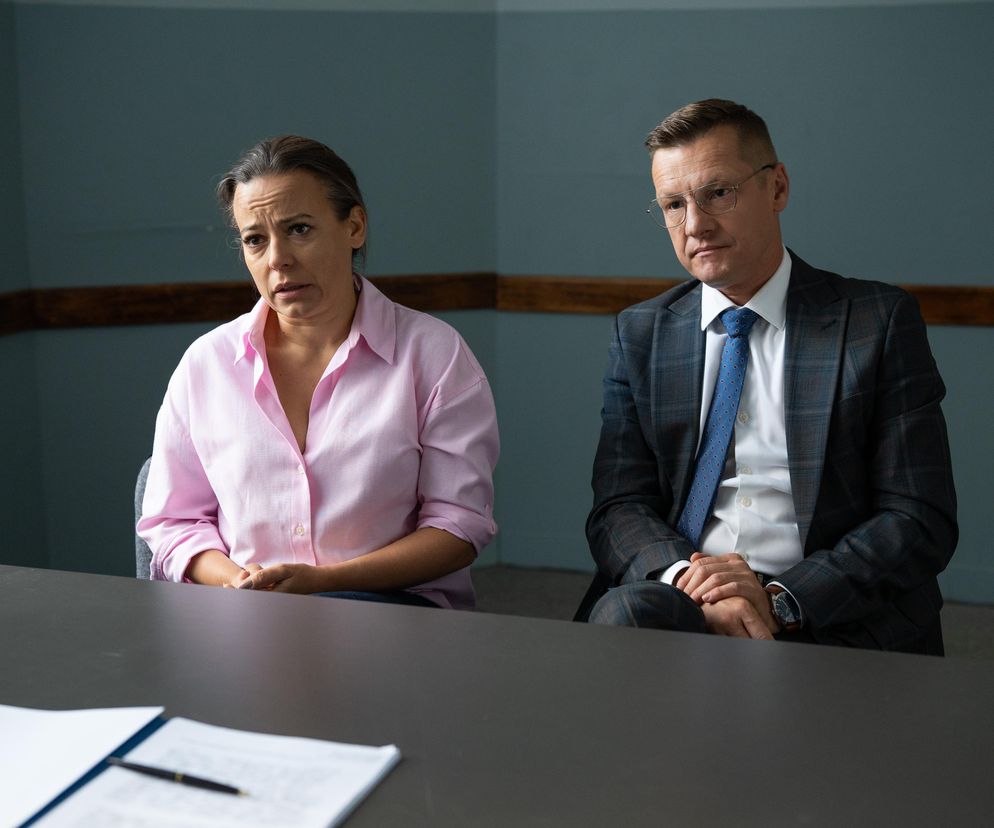 Zaniepokojona Magda Budzyńska (Anna Mucha) w różowej koszuli i zrezygnowany Piotrek Zduński (Marcin Mroczek) w eleganckim garniturze siedzą przy stole podczas przesłuchania, widoczne dokumenty i długopis. Więcej o ich losach w M jak miłość na portalu Super Seriale.