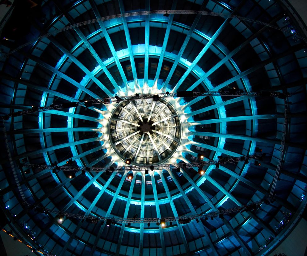 Kopuła Hali Stulecia we Wrocławiu, oświetlona błękitnymi i białymi światłami od wewnątrz, z widoczną kratownicą i centralnym świetlikiem. Fotografia ukazuje monumentalną konstrukcję żelbetową, ikonę architektury modernizmu. Więcej o tym dziele Maxa Berga przeczytasz na Architektura Murator Plus.