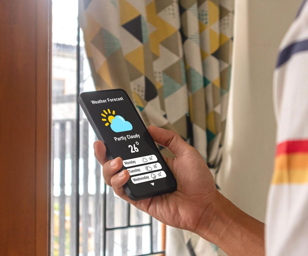 Osoba trzymająca smartfon z otwartą aplikacją Weather Forecast, pokazującą prognozę pogody na 26 stopni Celsjusza oraz na kolejne dni. Ekstremalne temperatury to ryzyko dla serca, o czym przeczytasz więcej na Poradnik Zdrowie.