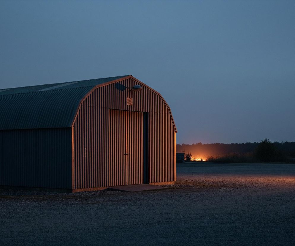 Blaszany budynek o zaokrąglonym dachu, przypominający hangar lub stodołę, z pionowymi panelami na ścianach. Fasada budynku, a zwłaszcza jego drzwi, jest oświetlona ciepłym, pomarańczowym światłem, które odbija się również na ciemnym, żwirowym podłożu przed nim. W tle, za budynkiem, widać ciemny las i intensywną, pomarańczową poświatę światła unoszącą się nad horyzontem, kontrastującą z ciemnym, granatowym niebem. Po prawej stronie, na pierwszym planie, smuga pomarańczowego światła rozciąga się w poprzek żwirowego terenu, co może sugerować obecność reflektorów.