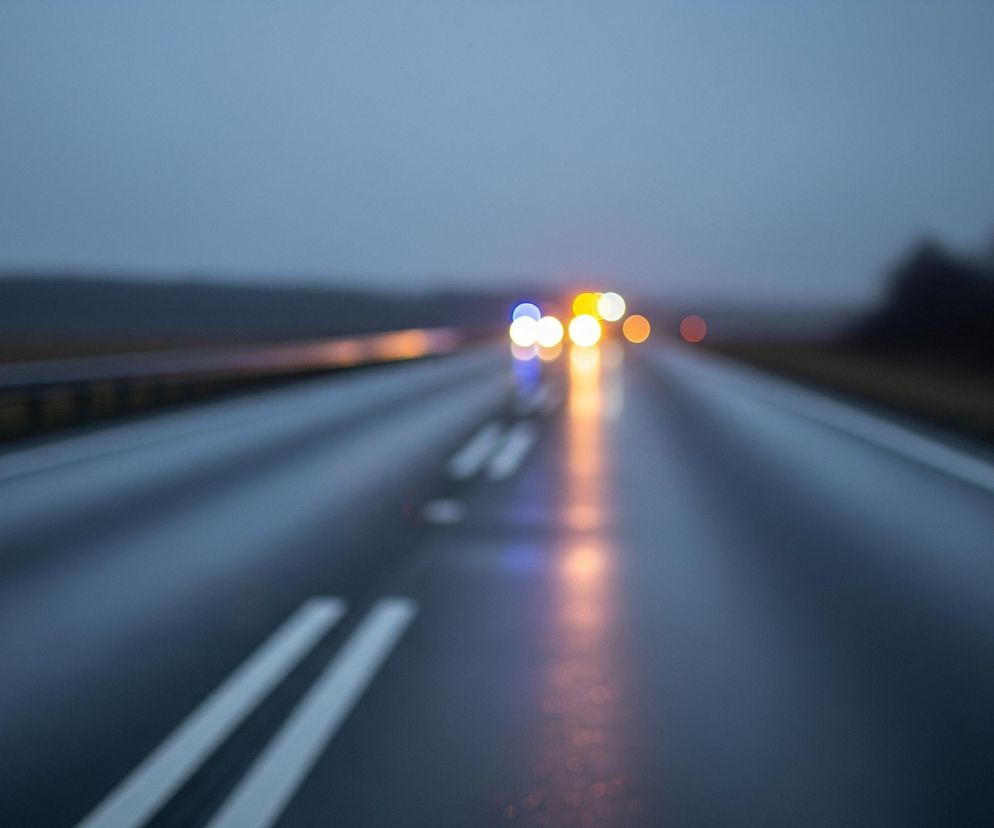 Rozmazaną drogę asfaltową w deszczową, pochmurną pogodę, która zanika w oddali. Na ciemnoszarej nawierzchni widoczne są odbicia kolorowych świateł, głównie żółtych, pomarańczowych i niebieskich, tworzących efekt bokeh. Po lewej stronie drogi biegnie metalowa barierka ochronna, a na horyzoncie majaczy niewyraźny zarys lasu.