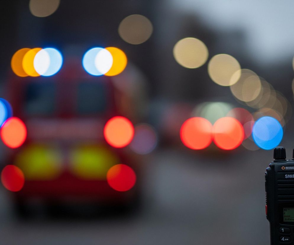 Na pierwszym planie, po prawej stronie, znajduje się czarne radiotelefon z anteną skierowaną lekko w lewo, z widocznym ekranem i klawiaturą numeryczną; na ekranie wyświetla się 100.0. Tło jest rozmyte, z widocznymi kolorowymi światłami typu bokeh, gdzie dominuje czerwień i pomarańcz, a także niebieskie i białe punkty świetlne. Czerwone światła w tle sugerują tył pojazdu ratunkowego, prawdopodobnie karetki lub wozu strażackiego, z migającymi niebieskimi światłami awaryjnymi.