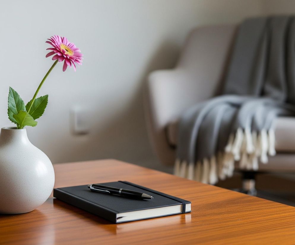 Na drewnianym stole stoi matowy, beżowy wazon z pojedynczą, różową gerberą, której żółty środek jest wyraźnie widoczny. Obok wazonu leży czarny notes z gumką i leżący na nim czarny długopis z metalowymi elementami. W tle, rozmyty, znajduje się beżowy fotel z narzuconym na niego szarym kocem zakończonym białymi frędzlami, a za nim widać jasną ścianę.