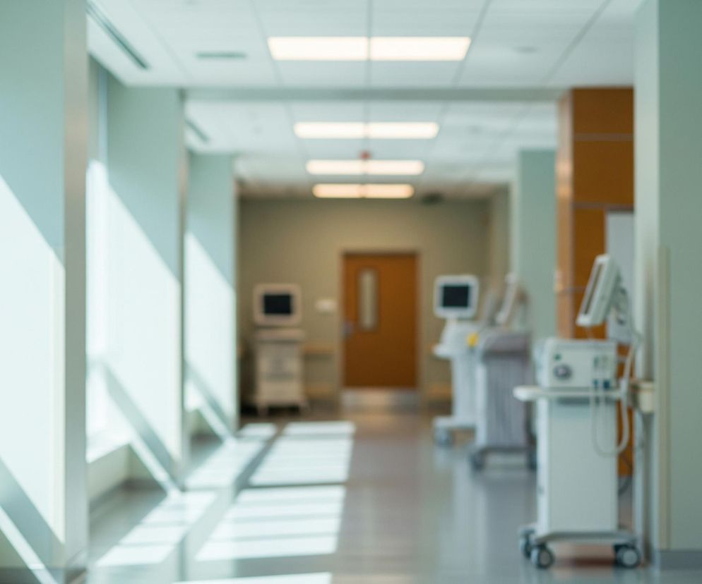 Jasny korytarz z wieloma oknami po lewej stronie dominuje w kadrze, z fragmentami światła słonecznego tworzącymi ukośne cienie na ścianie i podłodze. Po prawej stronie widać otwarte drzwi i białe urządzenie medyczne przymocowane do ściany. W oddali, korytarz prowadzi do niewyraźnych pomieszczeń z kolejnymi drzwiami i zamazanymi białymi wózkami medycznymi, każdy z ekranem u góry. Sufit korytarza jest oświetlony prostokątnymi lampami, a ściany mają jasnozielony odcień.