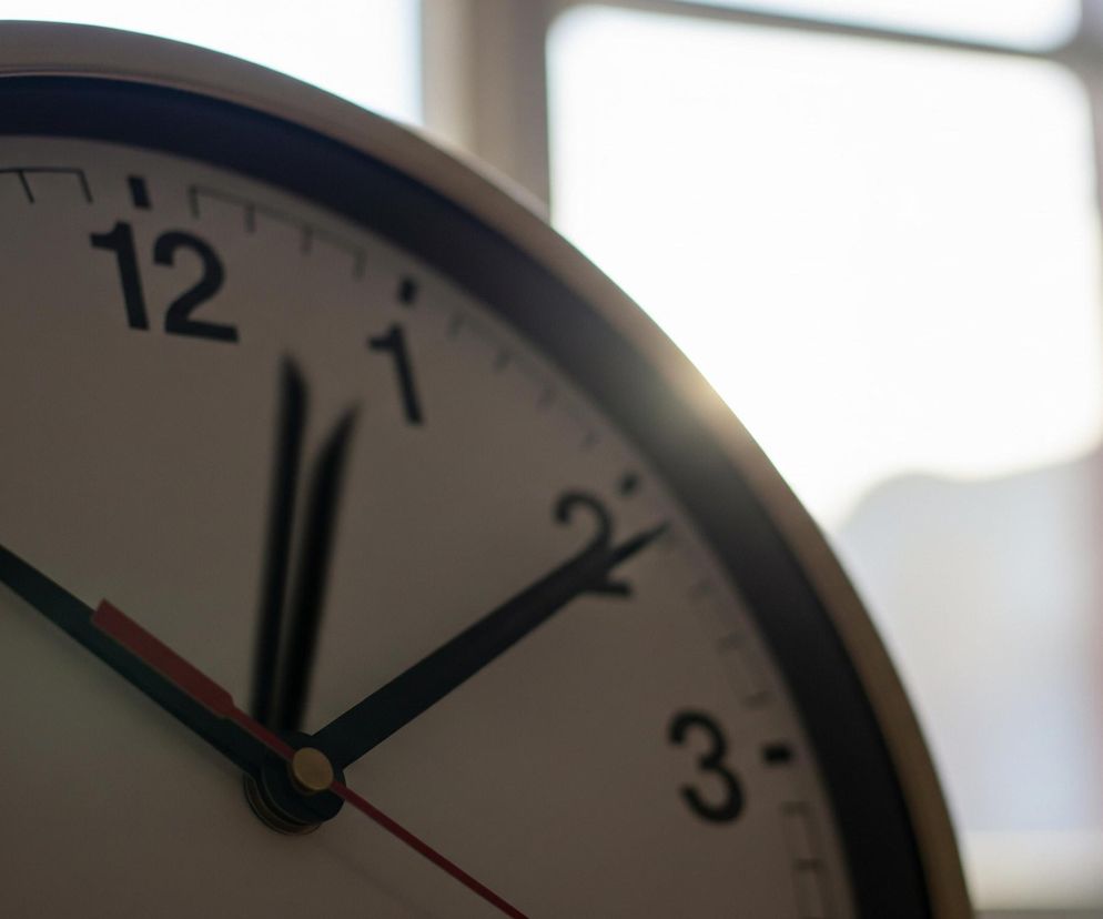 Zbliżenie na tarczę zegara ściennego, z jego górną prawą częścią rozświetloną jasnym, rozmazanym światłem pochodzącym zza obiektu. Tarcza jest biała z czarnymi cyframi od 9 do 3 i czarnymi kreskami minutowymi. Wskazówki są czarne, a czerwona, cieńsza wskazówka sekundowa, wychodzi z centralnego, złotego punktu. Zegar ma złotą ramkę.
