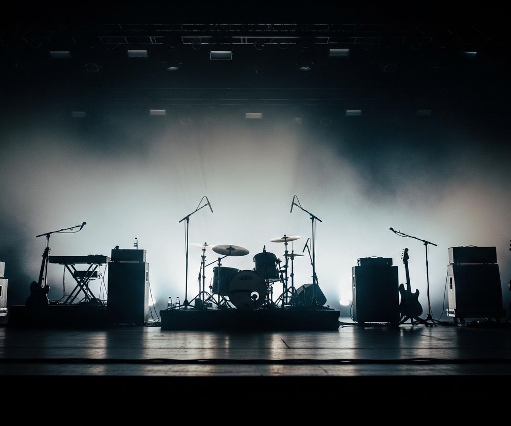 Na pierwszym planie widoczna jest scena, wypełniona sprzętem muzycznym. Centralnie umieszczona jest perkusja z widocznymi talerzami i bębnami, obok niej stoją mikrofony na statywach. Po lewej stronie perkusji znajduje się keyboard na statywie, gitara elektryczna oraz wzmacniacze, natomiast po prawej stronie kolejne wzmacniacze i dwie gitary, w tym jedna basowa. Całość oświetlona jest od tyłu mocnym, białym światłem, które tworzy dymny efekt i sprawia, że sprzęt jest w dużej mierze zacieniony. Górna część kadru i pierwszy plan pozostają w cieniu, co kontrastuje z jasnym tłem i podkreśla sylwetki instrumentów.