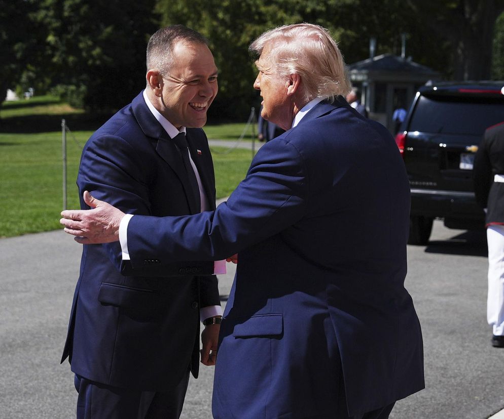 Uśmiechnięty Karol Nawrocki, ubrany w granatowy garnitur z białą koszulą i krawatem, wita się z Donaldem Trumpem, który ma na sobie podobny garnitur, obejmując go. Obaj panowie patrzą na siebie, a scena rozgrywa się na zewnątrz, z zielonym trawnikiem i czarnym samochodem w tle. Więcej informacji o spotkaniu przeczytasz na naszym portalu.