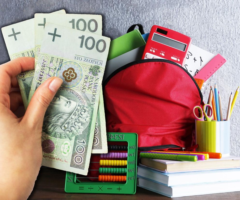 Ręka trzymająca banknoty stuzłotowe, obok czerwonego plecaka, książek i przyborów szkolnych.  Ilustracja do artykułu na Super Biznes o programie Dobry Start – 300 zł na wyprawkę szkolną.  Termin składania wniosków: do 31 sierpnia.