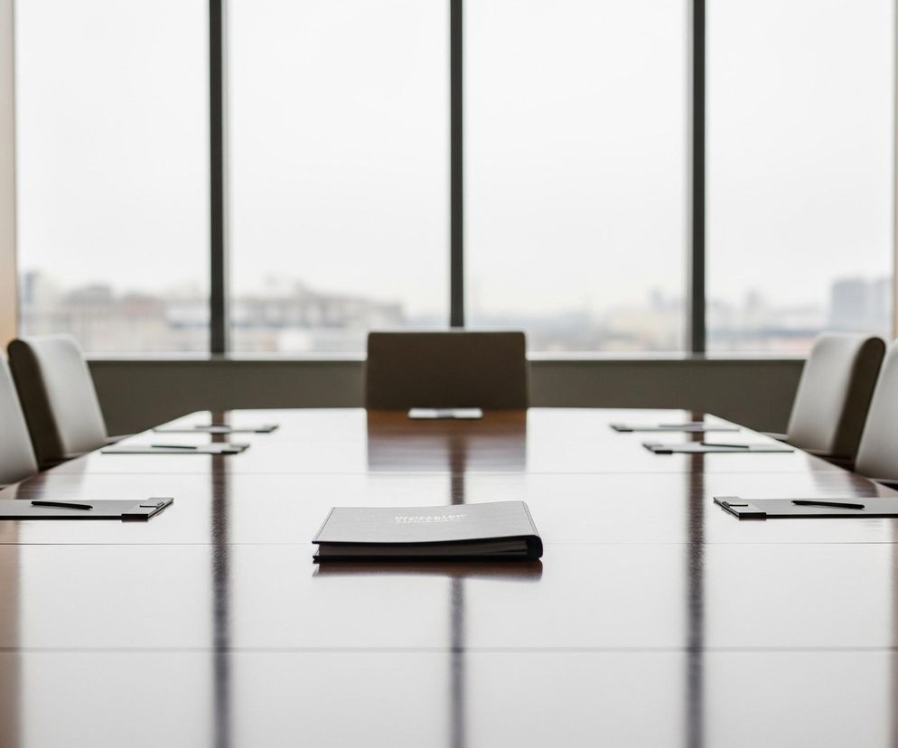 Długi, lśniący, drewniany stół konferencyjny w brązowym kolorze, zajmuje centrum kadru, rozciągając się od dołu obrazu. Na stole, wzdłuż jego boków, równomiernie rozmieszczono liczne jasnoszare podkładki pod notatki z ciemnymi długopisami, a na środku leży zamknięty, ciemny notes z wytłoczonym napisem. Wokół stołu ustawione są szare, skórzane krzesła z drewnianymi oparciami, widoczne częściowo, tworząc rząd po obu stronach. W tle znajduje się duże okno z pionowymi szprosami, przez które widać niewyraźny miejski krajobraz pod pochmurnym niebem, wpuszczające do pomieszczenia jasne, rozproszone światło.