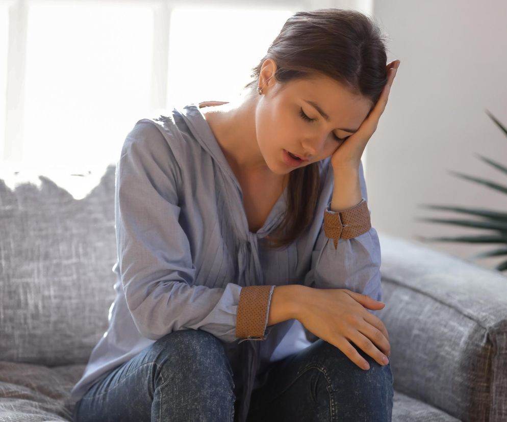 Kobieta siedząca na szarej kanapie, z zamkniętymi oczami i dłonią opartą o czoło, wygląda na zmęczoną, co symbolizuje skutki społeczne jet lagu i zmęczenia po zmianie czasu. O tym, jak sabotuje wagę i zwiększa ryzyko otyłości oraz cukrzycy, przeczytasz na Poradnik Zdrowie.