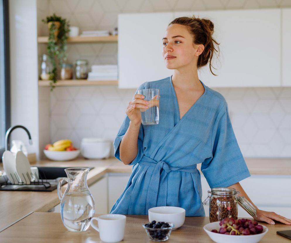 Młoda kobieta w niebieskim szlafroku pije wodę w kuchni, symbolizując poranne rytuały wzmacniające odporność. Obok niej dzbanek wody, miseczka z jagodami i winogronami oraz słoik z granolą. Artykuł na Poradnik Zdrowie podpowiada, jak zacząć dzień dla lepszego zdrowia.