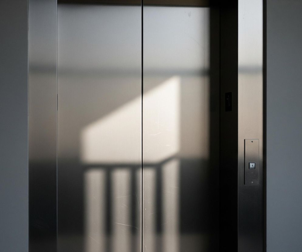 Dwoje zamkniętych, metalowych drzwi windy zajmuje centralną część kadru. Na ich błyszczącej, szarej powierzchni widoczne są odbicia światła i cieni, tworzące geometryczne kształty oraz fragmenty balustrady. Po prawej stronie drzwi znajduje się pionowy panel sterowania w kolorze ciemnoszarym z przyciskiem do wezwania windy, przypominającym kwadrat z białym trójkątem skierowanym w dół. Ściany po bokach drzwi są jednolicie ciemnoszare, stopniowo przechodzące w czerń na skrajach obrazu, co tworzy efekt głębi.