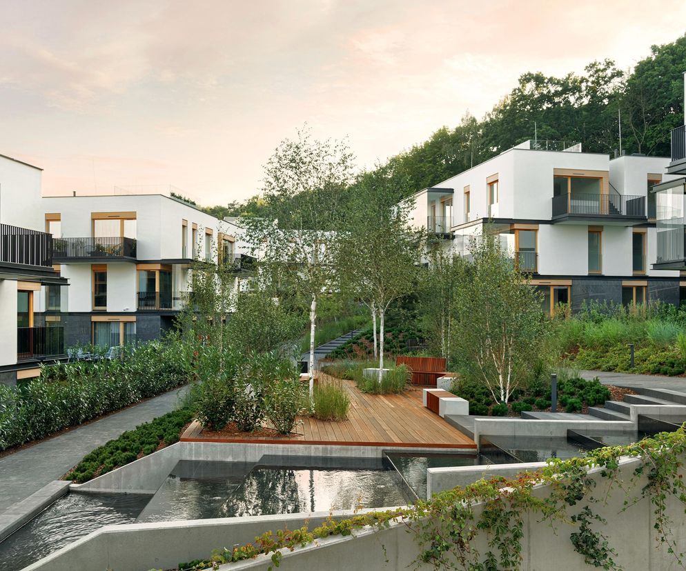 Białe, nowoczesne budynki mieszkalne z drewnianymi akcentami i balkonami z ciemnymi balustradami, wkomponowane w zielone otoczenie z drzewami, kaskadowymi zbiornikami wodnymi i drewnianymi tarasami. Jest to wizualizacja osiedla Nowe Kolibki w Gdyni, o którym przeczytasz na portalu Architektura Murator Plus.