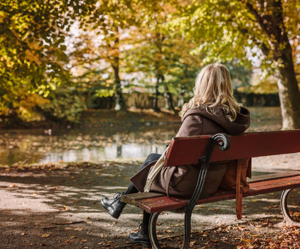 Kobieta z blond włosami, w brązowym płaszczu i szaliku, siedzi tyłem na czerwonej ławce, spoglądając na staw otoczony drzewami z jesiennymi liśćmi. Obraz symbolizuje samotność, poruszaną na Poradnik Zdrowie, zwłaszcza w kontekście pogorszenia samopoczucia jesienią.