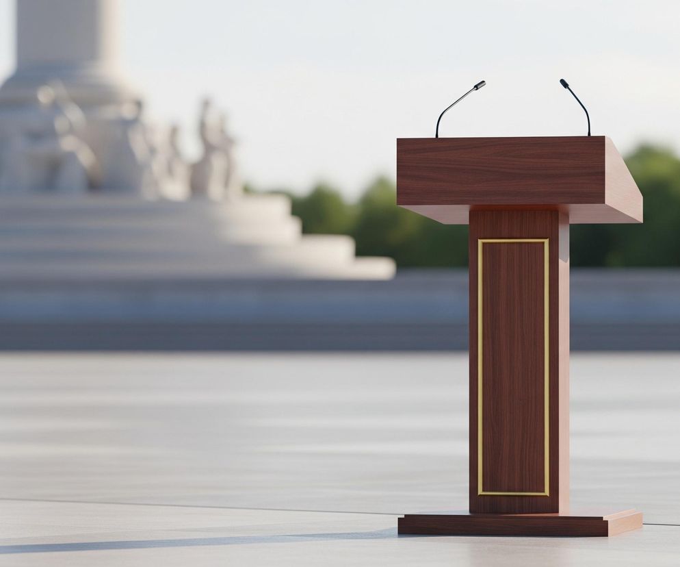 Na pierwszym planie, centralnie, stoi drewniana mównica o ciemnobrązowym kolorze, z prostymi, geometrycznymi kształtami. Mównica ma dwie mikrofony na szczycie, wychylające się do przodu na cienkich, czarnych statywach. Przednia część podstawy mównicy jest ozdobiona złotą ramką, tworzącą podłużny prostokąt. Mównica spoczywa na jasnej, płaskiej powierzchni, prawdopodobnie chodniku lub placu, oświetlonym jasnym światłem, rzucającym delikatne cienie.

W tle, po lewej stronie, rozmazany jest duży, monumentalny budynek lub pomnik z jasnego kamienia, z widocznymi schodami i fragmentami rzeźb, przypominających postacie ludzkie. Dalej, za budynkiem, widać pas zielonych drzew o niewyraźnych kształtach. Górna część obrazu to jasne, niemal białe niebo, wskazujące na dzień o słonecznej pogodzie.