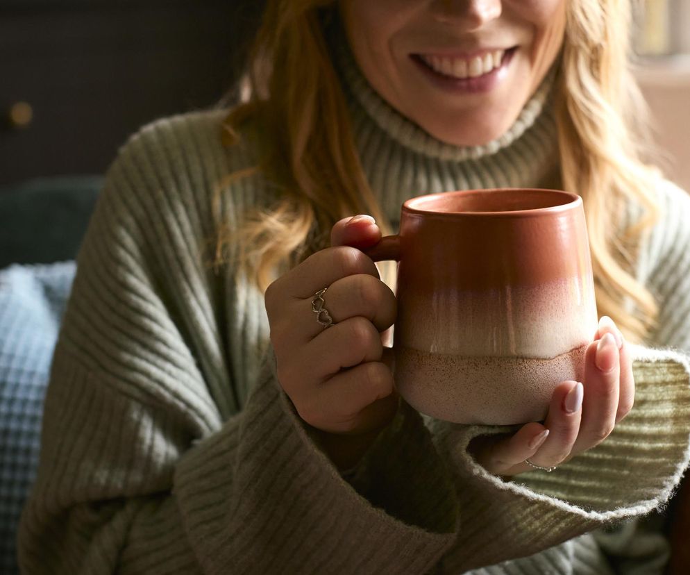 Uśmiechnięta blondynka w beżowym swetrze z golfem trzyma w dłoniach dwukolorowy ceramiczny kubek z gorącą herbatą. Na jej palcu widoczny jest złoty pierścionek z serduszkami. Więcej o właściwościach herbaty przeczytasz na Poradnik Zdrowie.