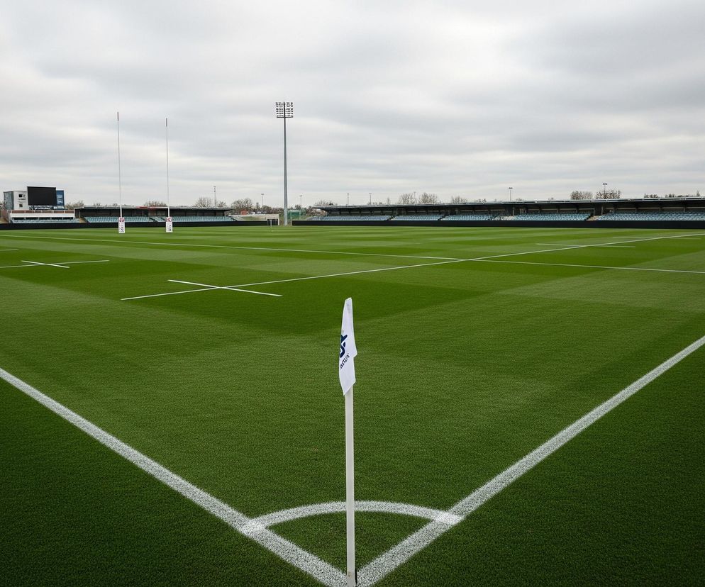 Kąt boiska do rugby z zieloną sztuczną trawą i białymi liniami, rozciągający się od prawego dolnego rogu ku górze. W prawym dolnym rogu stoi biała flaga z niebieskim logo na białej tyczce. Na tle boiska widać puste trybuny z niebieskimi siedzeniami, wysokie słupy z oświetleniem oraz budynki w oddali pod pochmurnym, szarym niebem.