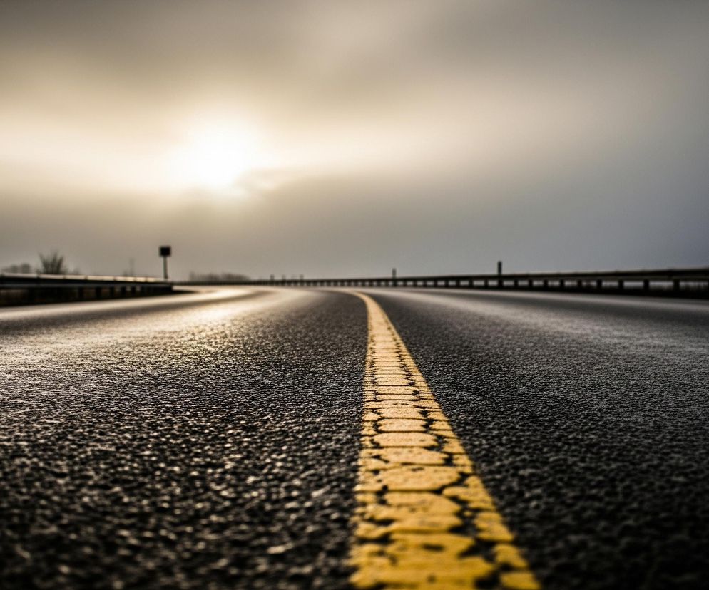 Czarna, asfaltowa droga biegnie przez środek kadru, z wyraźną, żółtą, przerywaną linią centralną, która oddala się w perspektywie. Nawierzchnia drogi jest mokra i błyszcząca, odbijając blade światło z górnej części nieba. Po obu stronach drogi widać ciemne barierki ochronne, które również znikają w oddali. Horyzont dominuje jasne, zamglone niebo z widocznym, intensywnym punktem świetlnym, sugerującym słońce, które rozjaśnia chmury.
