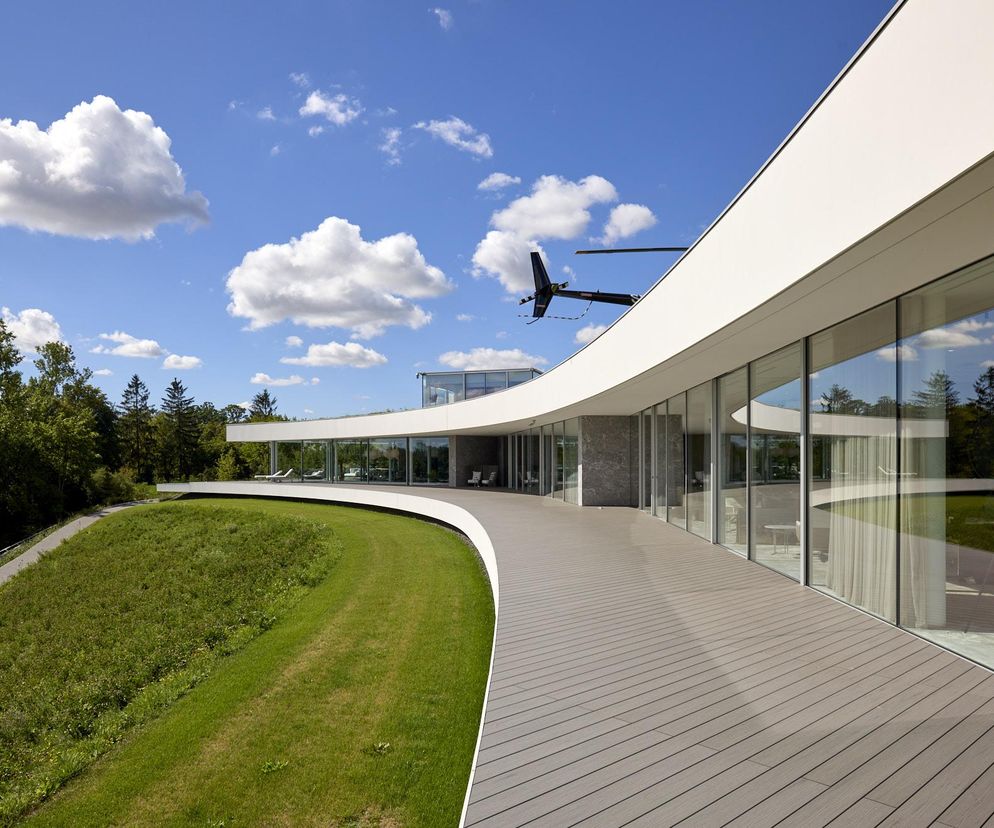 Nowoczesny, zakrzywiony budynek mieszkalny Dune House projektu Mobius Architekci, wpisujący się w krajobraz wzgórza nad Narwią, z obszernym tarasem i panoramicznymi oknami, otoczony zielenią i drzewami, pod błękitnym niebem z chmurami, a także unoszącym się helikopterem. Więcej o tej architekturze przeczytasz na Architektura Murator Plus.