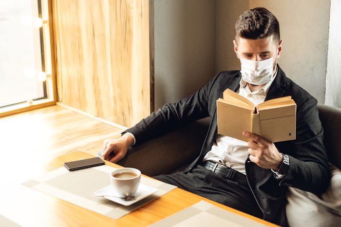 Mężczyzna w białej maseczce ochronnej i eleganckim stroju siedzi w restauracji, czytając książkę. Na stoliku przed nim znajduje się filiżanka kawy i smartfon. Zdjęcie ilustruje temat działania gastronomii w dobie koronawirusa. Więcej o restrykcjach przeczytasz na Poradnik Zdrowie.