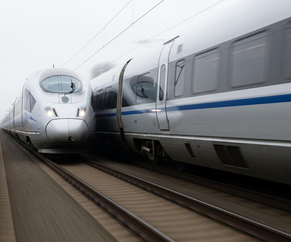 Dwa pociągi szybkobieżne, utrzymane w odcieniach szarości z niebieskimi akcentami, mijają się na równoległych torach pod pochmurnym niebem. Jeden pociąg, widoczny z przodu po lewej stronie, ma włączone światła, a jego aerodynamiczny kształt jest wyraźnie zarysowany. Drugi pociąg, zajmujący prawą część kadru, jest w ruchu, co widać po rozmytych liniach jego wagonów i szyb. Pasy torów i podłoża są rozmazane, co potęguje wrażenie prędkości.