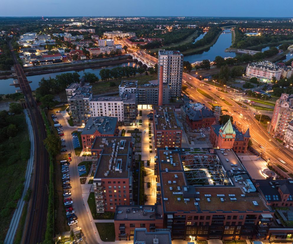 Osiedle Browary Wrocławskie z lotu ptaka o zmierzchu, z widocznymi ceglanymi budynkami, w tym zrewitalizowanym historycznym obiektem browaru i nowymi apartamentowcami, oświetlonymi uliczkami i rzeką Odrą w tle. Więcej o tym projekcie na Architektura Murator Plus.
