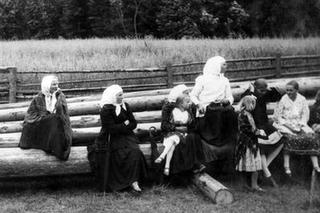 Kobiety w strojach regionalnych z dziećmi siedzą na drewnianych belkach.