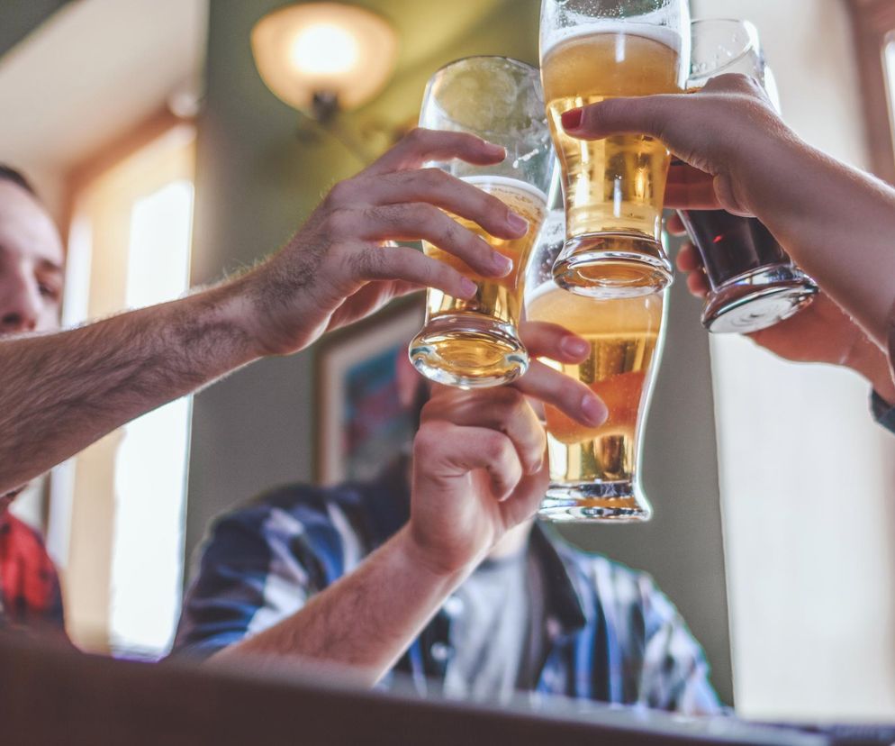 Grupa przyjaciół wznosi toast piwem w szklankach, celebrując wspólną chwilę. Dłoń z pomalowanymi paznokciami unosi ciemne piwo, podczas gdy pozostałe dłonie trzymają jasne piwo. Kontekst dotyczy wpływu nawet umiarkowanego spożycia alkoholu na zdrowie, o czym można przeczytać na portalu Poradnik Zdrowie.
