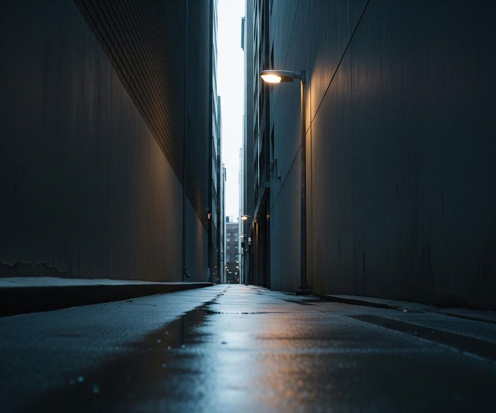 Wąska, ciemna uliczka lub przejście między wysokimi budynkami jest widoczne od poziomu ziemi. Powierzchnia ziemi jest mokra i błyszcząca, z wyraźnymi odbiciami świateł. Po prawej stronie, na ciemnej ścianie, zamocowana jest latarnia uliczna o ciepłym, pomarańczowym świetle, które rzuca smugę na ścianę i oświetla fragment mokrego chodnika. W tle, na końcu przejścia, widać więcej świateł i niebo.