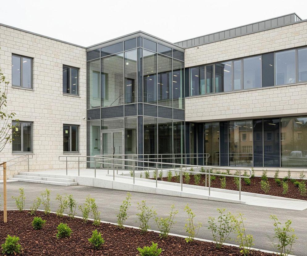 Nowoczesny dwupiętrowy budynek o elewacji z jasnobeżowej cegły dominuje na zdjęciu, z licznymi dużymi, prostokątnymi oknami o szarych ramach. Centralna część budynku to przeszklony wejście z podwójnymi drzwiami, otoczone również przeszklonymi ścianami. Przed wejściem znajduje się betonowa rampa dla osób niepełnosprawnych, zabezpieczona metalowymi poręczami, obok której widać schody. Wokół budynku rozpościerają się trawniki pokryte korą drzewną, w których rosną młode krzewy i drzewo z zielonymi liśćmi, wsparte na drewnianych palikach. Pochmurne, jasne niebo stanowi tło dla tej architektury.