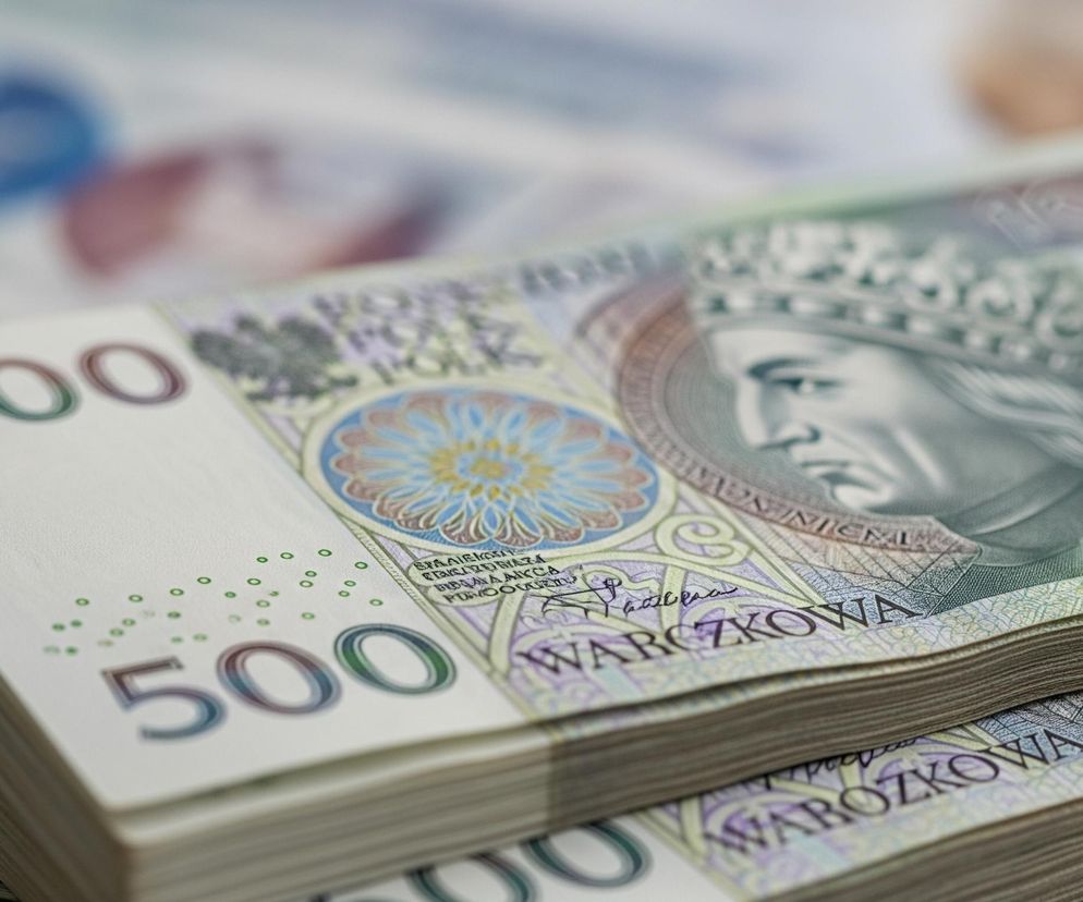 Duże zbliżenie przedstawia stos banknotów o nominale 500 złotych, ułożonych w dwa równoległe rzędy. Na wierzchu stosu widoczna jest jasna powierzchnia banknotu z nadrukowaną wartością 200, a pod nią widnieje wartość 500 w ciemniejszym, gradientowym kolorze. Banknoty zdobi portret króla w koronie, otoczony ozdobnymi wzorami w odcieniach zieleni, fioletu, błękitu i żółci, a także napisy POLSKIE i WARSZAWY. Tło jest rozmyte, ale można dostrzec inne banknoty w jasnych kolorach, z niewyraźnymi elementami wzorów.