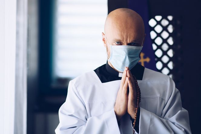 Łysy ksiądz w sutannie i maseczce ochronnej, z dłońmi złożonymi do modlitwy i różańcem, symbolizuje zmiany w uroczystościach religijnych w dobie pandemii. Więcej o organizacji Bożego Ciała przeczytasz na Poradnik Zdrowie.