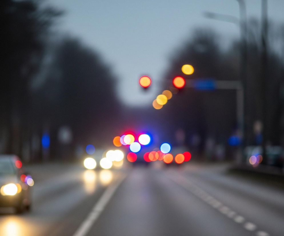 Głównym elementem obrazu jest szeroka droga asfaltowa widziana z perspektywy jadącego pojazdu, skupiona na scenie nocnej lub o zmierzchu. Po lewej stronie widoczny jest samochód z włączonymi, jasnymi reflektorami, które rozświetlają drogę przed nim. W oddali, na horyzoncie, rozmyte światła uliczne i sygnalizacji świetlnej tworzą kolorowe plamy – czerwone, pomarańczowe i niebieskie, sugerujące obecność innych pojazdów, być może z włączonymi światłami awaryjnymi lub sygnałowymi służb ratunkowych. Tło jest niewyraźne, z zarysem drzew i budynków po obu stronach drogi, co potęguje wrażenie ruchu i skupienia na świetlistych punktach.