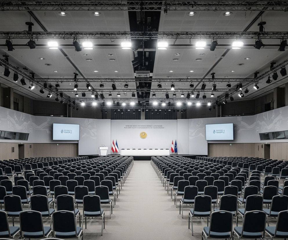 Obszerne, puste audytorium z rzędami krzeseł zajmuje dużą część pomieszczenia. Krzesła, z granatowymi obiciami i metalowymi ramami, ustawione są symetrycznie, tworząc szerokie przejście w centralnej części. Na wprost znajduje się podwyższona scena z białym stołem i mównicą, po której bokach stoją flagi, w tym biało-czerwono-biała i niebieska z gwiazdami. Na ścianie za sceną widoczne są dwa duże ekrany prezentujące grafikę, a pomiędzy nimi tablica z tekstem w obcym języku i złotym herbem. Sufit pokrywa skomplikowana konstrukcja z metalowych kratownic i reflektorów oświetlających całe wnętrze.