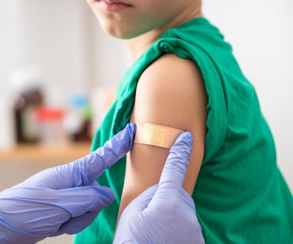 Dłoń osoby w fioletowej rękawiczce medycznej przyklejająca plaster na ramię dziecka po szczepieniu. Dziecko ma na sobie zieloną koszulkę. Zdjęcie ilustruje zmiany w zasadach rozliczania szczepień w aptekach, o których można przeczytać na portalu Poradnik Zdrowie.