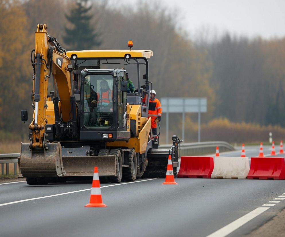 Na pierwszym planie, z lewej strony, widnieje pomarańczowo-biały pachołek drogowy. Centralną część zdjęcia zajmuje żółta koparka kołowa z operatorem w pomarańczowej kamizelce odblaskowej w kabinie. Za koparką, po prawej stronie drogi, ustawiony jest rząd pomarańczowych i białych barier drogowych, za którymi w oddali widać szereg pomarańczowych pachołków. W tle rozciąga się pasmo drzew o jesiennych, żółto-pomarańczowych liściach, a nad nimi szare, zachmurzone niebo. Po prawej stronie drogi, tuż za barierami, stoi słup z okrągłym znakiem drogowym o czerwonej obwódce i symbolu mostu zwodzonego, a pod nim prostokątny niebieski znak ze strzałką w prawo.