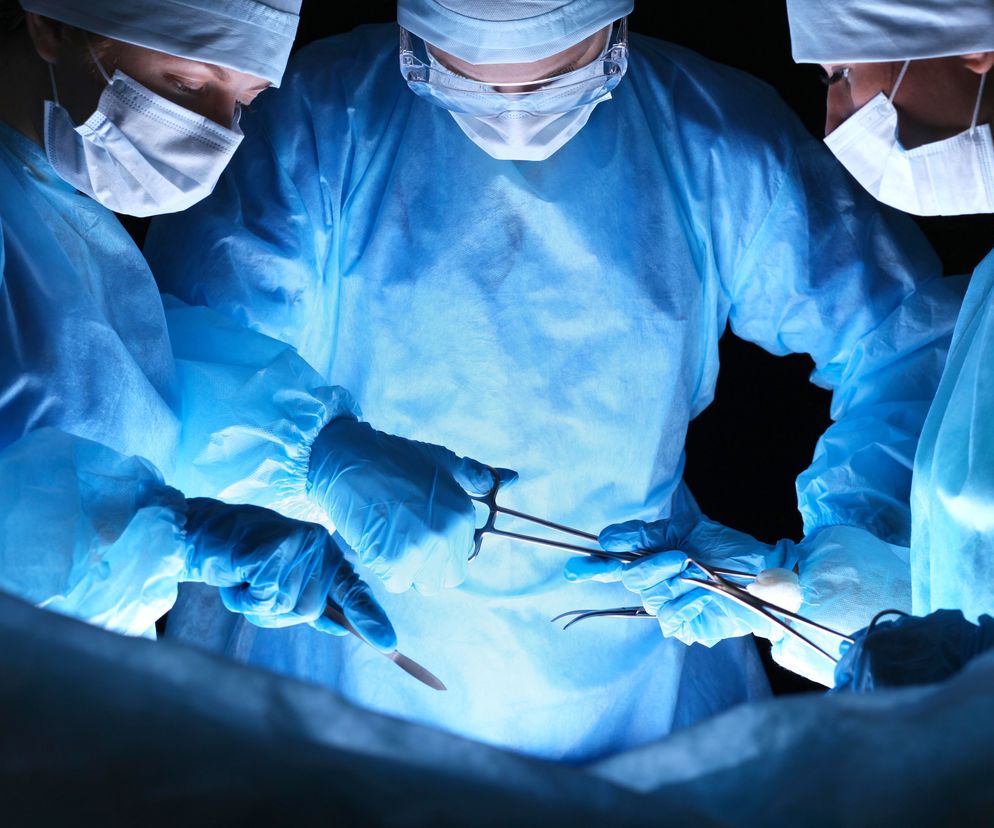 Zespół trzech chirurgów w niebieskich fartuchach i maskach, wykonujących operację z użyciem narzędzi medycznych, oświetlonych jasnym, niebieskim światłem. Temat innowacyjnych metod w kardiochirurgii, jak CorMatrix, można znaleźć na Poradnik Zdrowie.