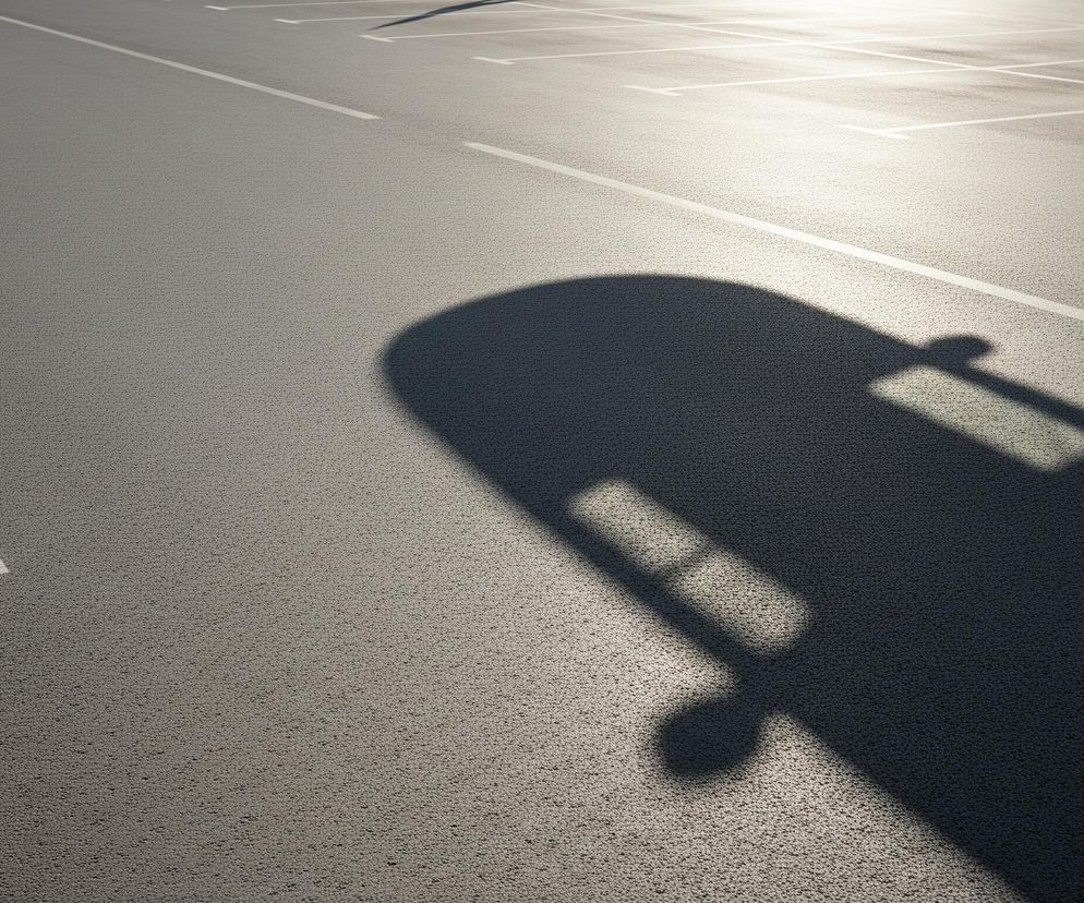 Na ciemnoszarym, nierównym asfalcie parkingu widoczny jest wyraźny, ciemny cień samochodu, rzucany z prawej strony kadru. Na nawierzchni są białe linie wyznaczające miejsca parkingowe, z literami 'T' tworzącymi ramki. Lewa część parkingu jest zacieniona, natomiast prawa strona jest jasno oświetlona przez słońce, co tworzy wyraźny kontrast.