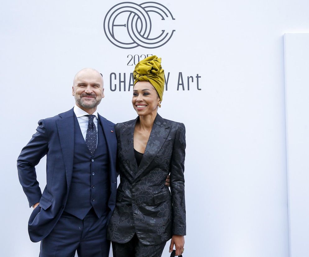 Rafał Brzoska w eleganckim garniturze i Omenaa Mensah w kwiecistym żakiecie i turbanie, pozują na tle białej ściany z logo gali charytatywnej. Oboje uśmiechają się. O ich działalności można przeczytać na Super Biznes.